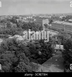 Un'ampia fotografia panoramica in bianco e nero degli anni '1980, che mostra il paesaggio urbano di Sloviansk, RSS Ucraina, da un alto punto panoramico. La vista cattura l'area intorno a Svobody Street, presentando una varietà di arazzi urbani caratteristici dell'era sovietica. Il paesaggio e' un mix di blocchi di appartamenti a pannelli multipiano, vecchi edifici amministrativi e una estesa e bassa area residenziale con case private annidate tra lussureggiante vegetazione. In lontananza, uno skyline industriale con camini di fabbrica affonda saldamente la città nel cuore industriale del Donbas Foto Stock