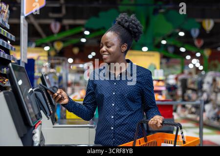 Giovane donna nera che esegue la scansione della spesa e paga con uno smartphone al supermercato, sorridendo mentre utilizza nfc contactless per fare acquisti al dettaglio rapidi e convenienti Foto Stock