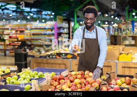 Assistente di negozio maschile che indossa un grembiule e guanti, in piedi in un supermercato produce Aisle e tiene felicemente in mano una mela rossa fresca, garantendo qualità ai clienti Foto Stock