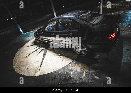 Roma, italia 30 settembre 2025: Berlina bmw e46 nera parcheggiata su cemento bagnato sotto un riflettore circolare di notte, riflessi lucidi e atmosfera urbana Foto Stock