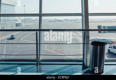 Un terminal aeroportuale vuoto con grandi finestre che si affacciano sulla pista. Un cestino è visibile in primo piano. La scena è luminosa e spaziosa. Foto Stock