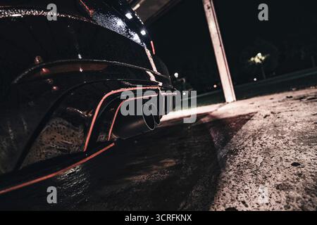 Roma, italia 30 settembre 2025: Paraurti posteriore nero per auto sportive e doppi scarichi scintillanti con gocce di pioggia, illuminati di notte in una strada urbana vuota Foto Stock