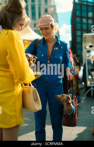 Londra, Regno Unito – 2025.09.27 – Una donna con una tuta blu e un berretto rosa porta due dachshunds, uno nella sua borsa, mentre chiacchiera con un'altra donna al CLA Foto Stock