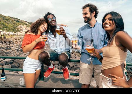 Un gruppo molto felice di giovani e diversi amici condivide una gioia, abbracciando e bevendo birra artigianale insieme in riva al mare durante una splendida holi estiva Foto Stock