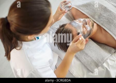 Vista dall'alto dell'estetista che applica la maschera cosmetica in argilla con un pennello sul viso dell'uomo nel salone di bellezza Foto Stock