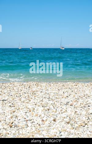 spiaggia di ciottoli bianchi alla luce del sole sul mare e sul cielo all'alba. Yacht all'orizzonte Foto Stock