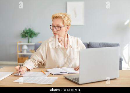 Donna anziana a casa che scrive appunti, usa un laptop, siede a tavola Foto Stock
