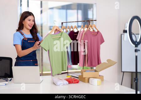 Happy Asian Woman promuove la nuova collezione di abbigliamento tramite il live streaming dal suo luminoso e moderno studio che gestisce il packaging degli ordini online su misura Foto Stock