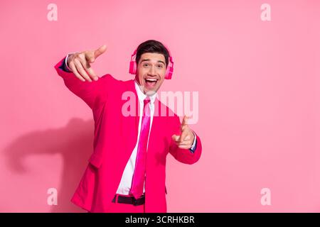 Giovane energico con un abito rosa brillante e cuffie che ascolta musica e posa elegante su uno sfondo rosa Foto Stock