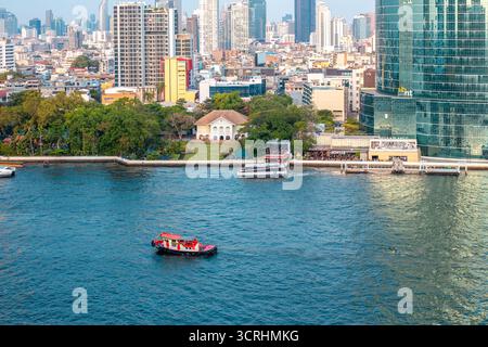 Lungo un ampio fiume, pieno di barche e chiatte sotto un cielo luminoso, si dispiega un vasto paesaggio urbano che mostra una miscela di architettura moderna e tradizionale. Bangkok, Thailandia - 26 marzo 2024 Foto Stock