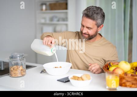 L'uomo 40 fa colazione in cucina. Il ragazzo ama il cibo fresco al tavolo della cucina la mattina. Un uomo mangia cereali e frutta in cucina. Uomo Foto Stock