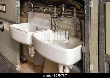 Lavandini industriali doppi in un bagno pubblico usurato con tubi a vista e rubinetti cromati Foto Stock