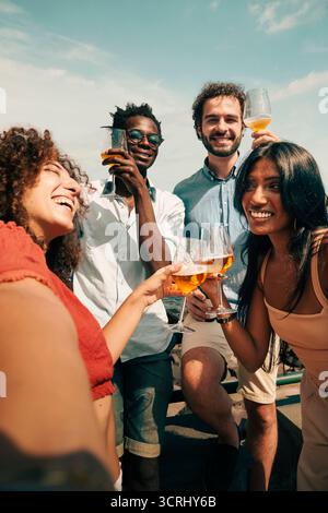 Un allegro gruppo multiculturale di amici che fanno un divertente selfie POV mentre brinano con bicchieri di birra artigianale, celebrano una vacanza estiva insieme all'aperto Foto Stock