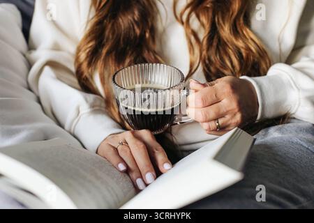 Donna che tiene la tazza di caffè a mano. Momenti di vita con una bevanda calda. Libro da lettura all'interno con dettagli accoglienti. Concetto di relax con atmosfera informale. Foto Stock