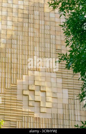 Dettaglio della facciata di un edificio moderno caratterizzato da piastrelle geometriche con motivi rialzati e incassati, parzialmente incorniciate da foglie verdi in parte Foto Stock
