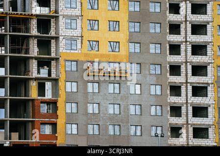Building facade undergoing insulation installation. Construction workers on scaffolding working on residential skyscraper exterior. Modern multi-story apartment block development in urban environment Foto Stock