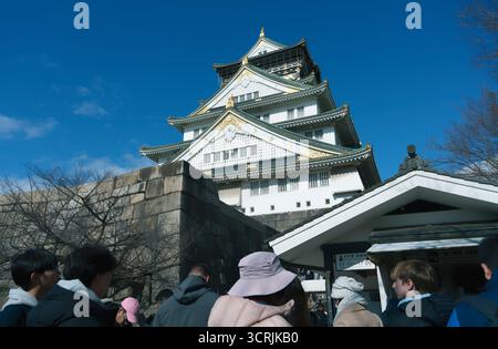I visitatori fanno la fila all'ingresso del Castello di Osaka, con facciata bianca e dettagli dorati che brillano sotto il cielo azzurro Foto Stock