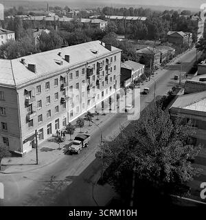Una fotografia d'archivio in bianco e nero ad alto angolo del 1988, che cattura una scena di strada in via Yunykh Kommunarov a Sloviansk, RSS Ucraina. La vista è centrata su un grande edificio residenziale a 4 piani, un classico esempio di architettura neoclassica sovietica del dopoguerra. Sotto, la strada mostra il traffico giornaliero tipico dell'epoca, tra cui un classico camion sovietico GAZ e diverse auto. Questo paesaggio urbano panoramico offre uno sguardo autentico e dettagliato alla vita quotidiana e all'ambiente urbano di una città provinciale durante il periodo Perestroika nella tarda URSS Foto Stock