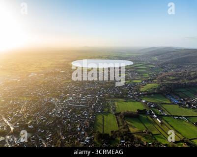 Vista aerea della luce dorata del sole che baciano i campi verdi e i tetti e il sereno bacino idrico che rispecchia il cielo, Cheddar, Inghilterra, Regno Unito. Foto Stock