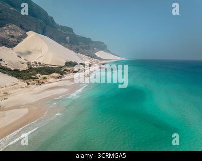 Vista aerea delle onde turchesi si adagia dolcemente contro la spiaggia sabbiosa sotto le torreggianti scogliere e le aspre dune bianche di Socotra, Socotra, Socotra, Yemen. Foto Stock