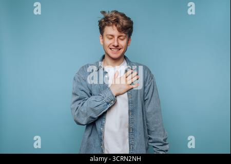 Un giovane sorridente in piedi su uno sfondo blu con una mano sul petto esprimendo gioia e gratitudine Foto Stock