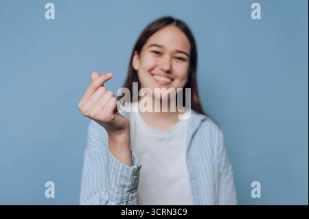 Una giovane donna gioiosa mostra un gesto cardiaco con le dita, in piedi davanti a uno sfondo azzurro. Il suo sorriso irradia felicità e positivit Foto Stock