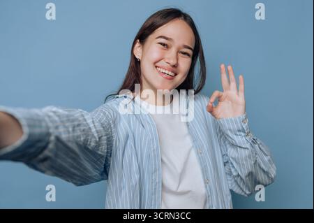Una giovane donna sta scattando con gioia un selfie. Indossa una camicia a righe e fa un gesto "okay" alla mano. Lo sfondo azzurro migliora il Foto Stock