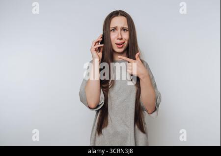 Una giovane donna con i capelli lunghi si staglia su uno sfondo semplice, sembra sorpresa mentre parla al telefono e si rivolge verso se stessa, trasmettendo un senso Foto Stock