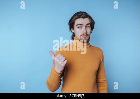 Giovane uomo con dolcevita senape che gesta con il pollice su sfondo blu. Foto Stock