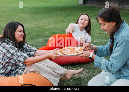 Amici che si rilassano su sacchi di fagioli colorati all'aperto, condividendo cibo e bevande in momenti di vita informale di incontro sociale con un cane piccolo. Donna informale Foto Stock