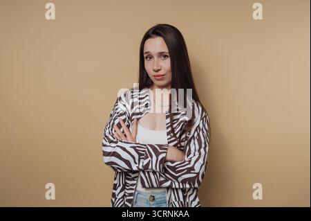 Giovane donna con capelli lunghi in una camicia a motivi zebrati, braccia incrociate, sfondo neutro. Foto Stock