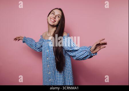 Una donna gioiosa con un abito a scacchi blu si posa su uno sfondo rosa, sorridendo e allungando le braccia giocosamente. Foto Stock