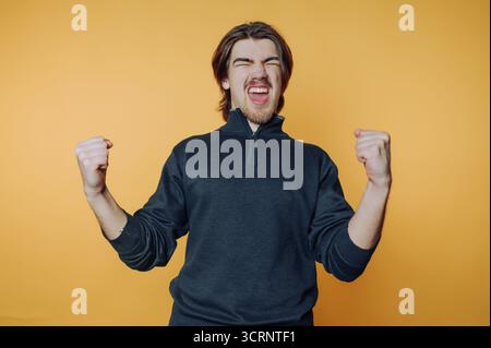 ritratto di giovane uomo alza il pugno di entusiasmo, trasmettendo un senso di celebrazione e vittoria su sfondo giallo Foto Stock
