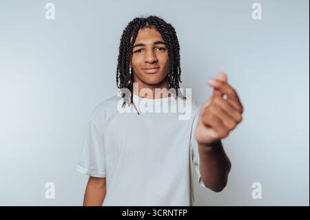 ritratto di un giovane uomo guarda scherzosamente la fotocamera mentre forma un cuore con le dita, irradiando calore e affetto su sfondo chiaro Foto Stock