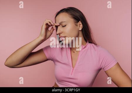 Una giovane donna vestita con una camicia rosa si erge su un morbido sfondo rosa, visibilmente stressata mentre si appoggia la mano contro la fronte, esprimendo d Foto Stock
