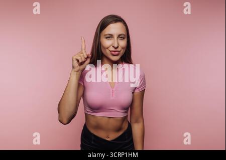 Una donna sta su un solido sfondo rosa, sorridendo e giocherellando alzando il dito. Trasuda fiducia e disponibilità nella sua fuga informale Foto Stock