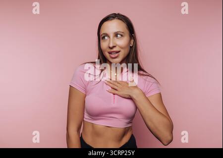 Una giovane donna si erge con sicurezza su uno sfondo rosa, sorridendo e mettendo una mano sul petto. Irradia energia e giocosità nella sua a casual Foto Stock