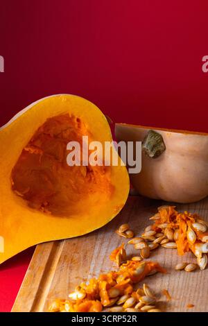 Zucca fresca tagliata a metà esposta su un tagliere di legno con semi e polpa sparsi intorno, su un audace sfondo rosso Foto Stock