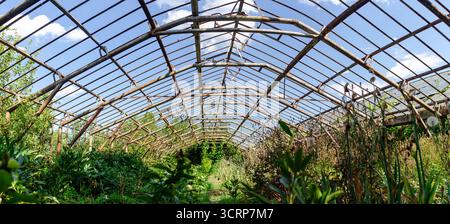 Serra vuota abbandonata con struttura metallica e tubi arrugginiti, finestre rotte attraverso le quali nuvole bianche sono visibili nel cielo blu. La serra è troppo grande Foto Stock