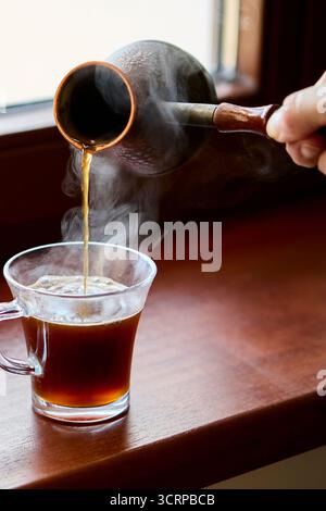 Una tazza di vetro trasparente fumante riempita di caffè nero si trova su un davanzale in legno. Il vapore si innalza di luce naturale creando un ambiente caldo e confortevole. Foto Stock