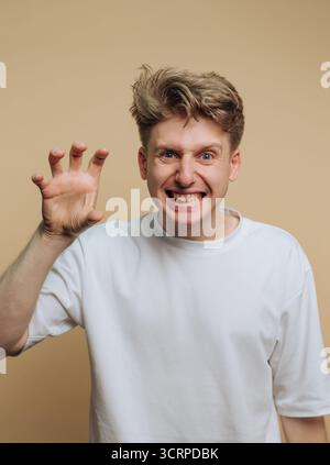 Un giovane con i capelli biondi duri sorride molto mentre imita giocosamente un gesto di artiglio con la mano. Lo sfondo neutro mette in risalto il suo cheerf Foto Stock