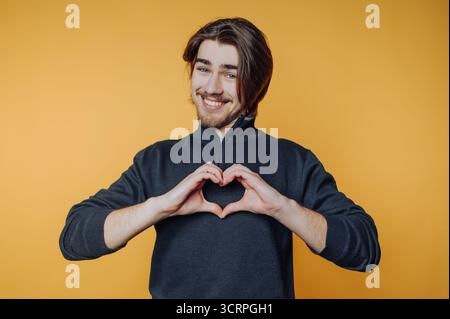 ritratto di un giovane uomo fa un gesto cardiaco con le mani su sfondo giallo, ritraendo gentilezza e amore. Foto Stock