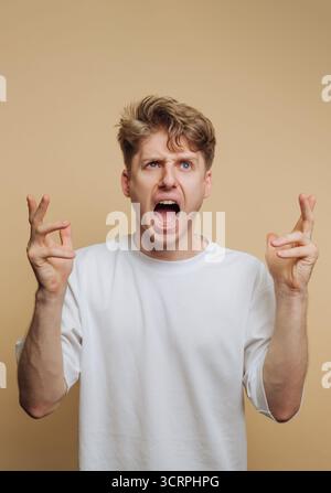 ritratto di un giovane che esprime speranza e anticipazione con le dita incrociate, con un'espressione esuberante su sfondo beige Foto Stock