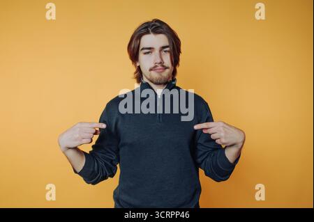 Giovane con un maglione scuro che punta su se stesso su uno sfondo arancione. Foto Stock