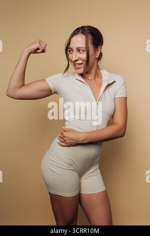 Una donna si trova con fiducia in un vestito beige, flessendo i muscoli del braccio. Sorride calorosamente alla telecamera, incarnando l'empowerment e il fitness in un semplice Foto Stock