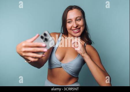 Una giovane donna che indossa un top azzurro sorride mentre si fa un selfie. Le piace il momento, posare giocherellamente davanti a uno sfondo blu fisso. Foto Stock