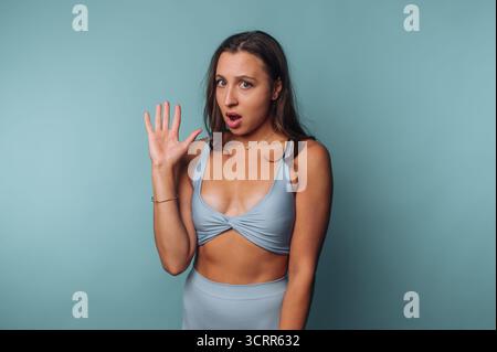 Una giovane donna si staglia su uno sfondo blu pieno, indossando un top in cotone blu e un fondo a vita alta. Alza una mano, mostrando un ex sorpreso Foto Stock