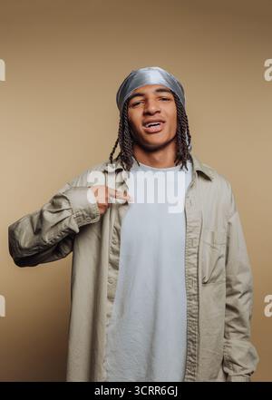 Un giovane si comporta in modo rilassato, sorridendo leggermente mentre indica la sua camicia. Il suo velo argentato completa il suo outfit leggero, creando Foto Stock