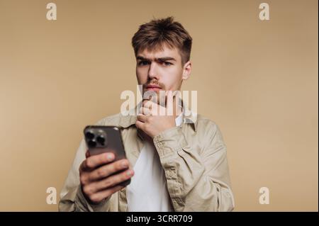 Un giovane uomo sta riflettendo su qualcosa di importante quando tiene uno smartphone in una mano. Ha un'espressione premurosa ed è vestito in modo informale, in piedi Foto Stock