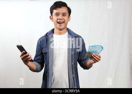 Un giovane sorridente in un abbigliamento casual tiene uno smartphone in una mano e una pila di banconote della Rupia indonesiana Foto Stock